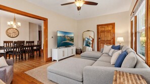 A living room with a sectional sofa, a dining area with wooden chairs, a TV on a white cabinet, and a large mirror on the wall. - DT Libertyville Home w/ Arcade PacMan by Navy Base (Libertyville)