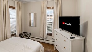 Een slaapkamer met een bed, een televisie op een dressoir, een plafondventilator en ramen voorzien van gordijnen.
