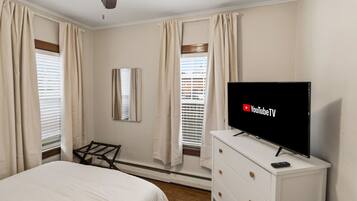 A bedroom with a bed, a TV on a dresser, a ceiling fan, and windows with curtains.