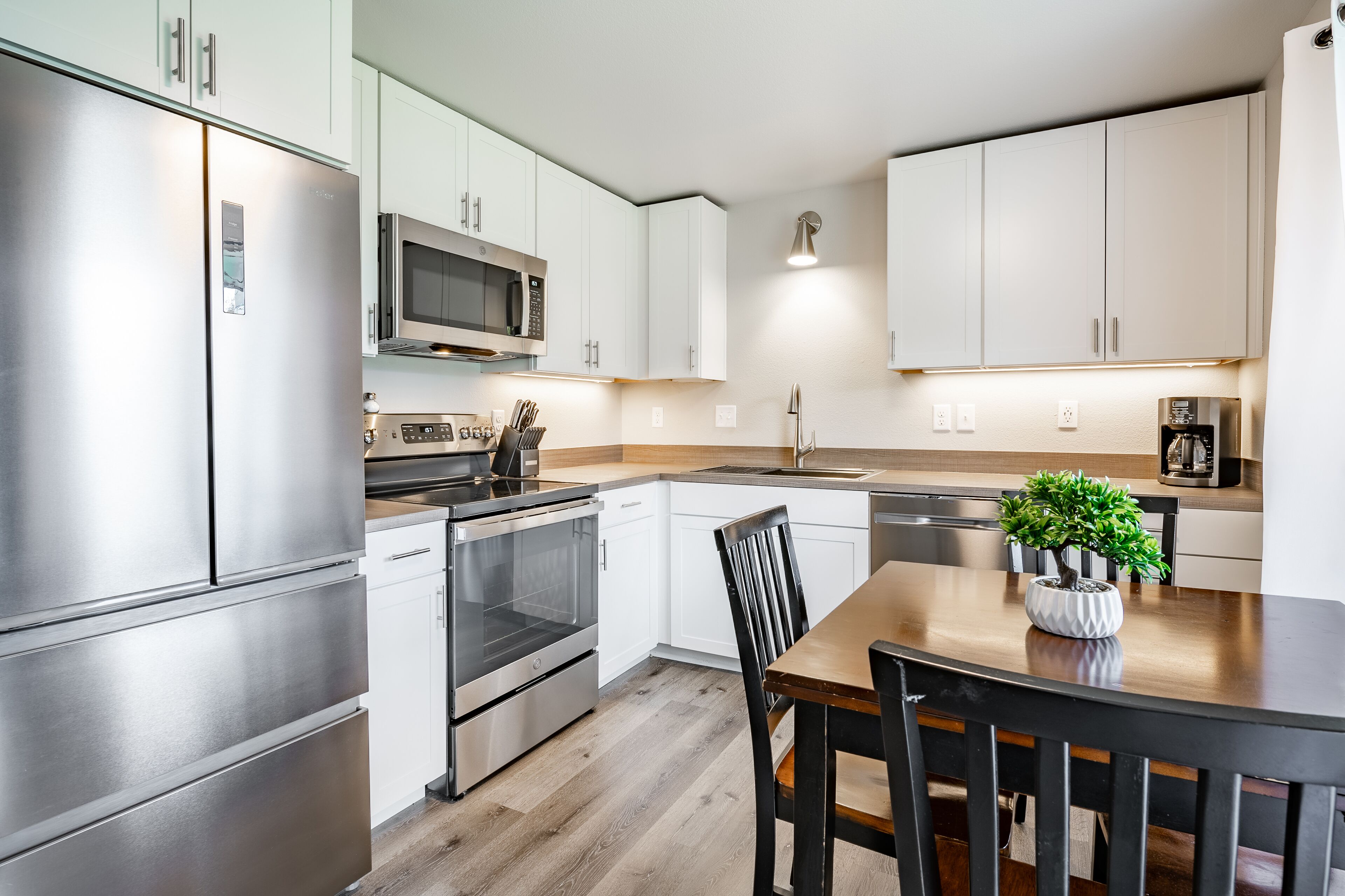 A modern kitchen with stainless steel appliances, white cabinetry, and a wooden dining table with a potted plant.