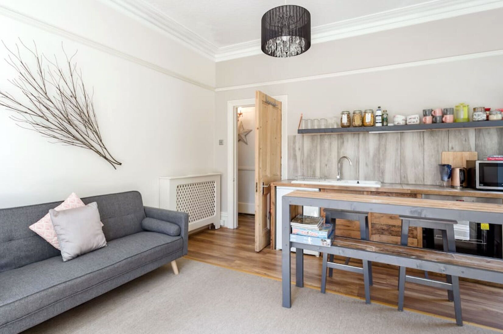 Una sala moderna con un sofá gris, una mesa de comedor de madera y una zona de cocina con un fregadero y estanterías.