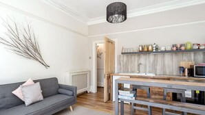 A modern living room with a grey sofa, a wooden dining table, and a kitchen area with a sink and shelves. - Rustic Revived Victorian Seaside Retreat (Bournemouth)