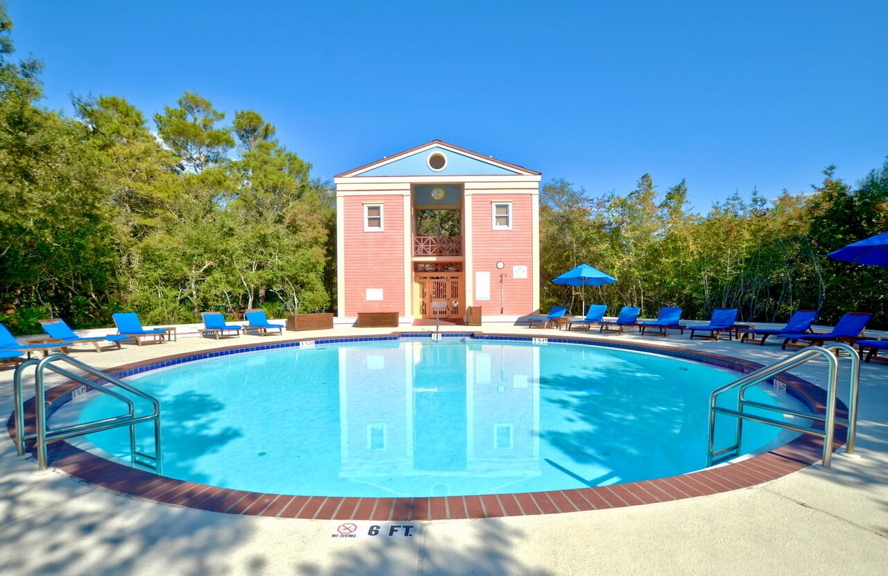A swimming pool with a red building in the background, surrounded by blue lounge chairs and umbrellas.