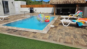 A swimming pool with a thatched-roof structure, surrounded by white lounge chairs and a grassy area. - Aruba Personal Paradise-2BR Home w/ private pool, convenient location (Noord)