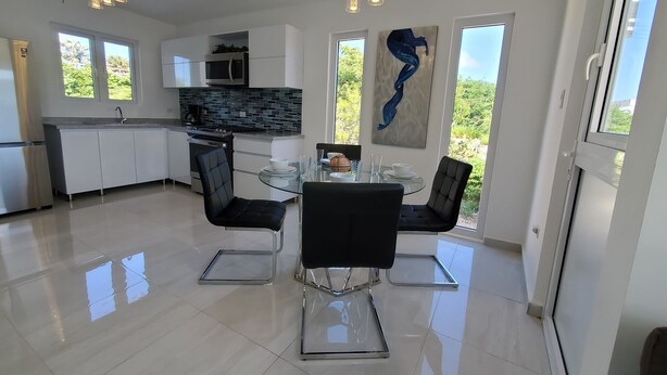 A modern kitchen with white cabinets, a black countertop, and a stainless steel refrigerator. There is a dining area with a glass table and black chairs. - Aruba Personal Paradise-2BR Home w/ private pool, convenient location (Noord)