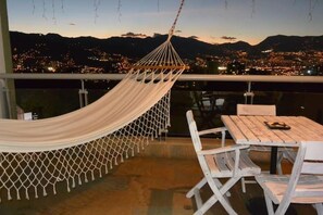 Un hamac sur un balcon, avec une table et des chaises, d’où l’on peut admirer la vue sur la ville au crépuscule.