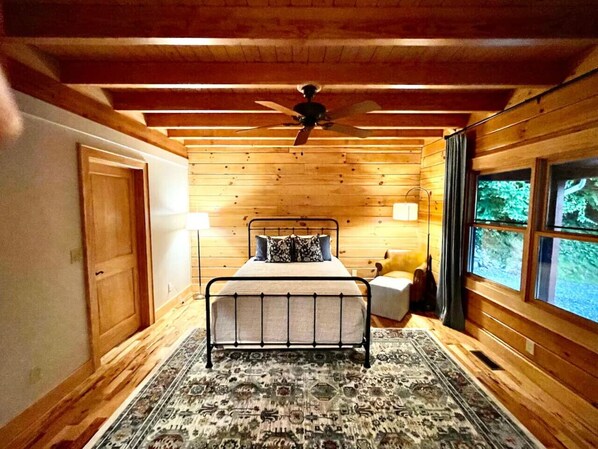 A bedroom with a wooden ceiling, a bed with a headboard, a bedside table, a lamp, and a window with curtains.