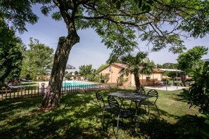 Un espace piscine avec une terrasse entourée d’une clôture, une table avec des chaises, et un bâtiment avec un toit rouge en arrière-plan.