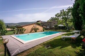 A swimming pool with a tiled deck, a grassy area with lounge chairs, and a building with a red-tiled roof. - Villa Gelsi by MMega (Cortona)