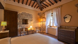 Een slaapkamer met een bed, een dressoir, een stenen muur, een lamp, een stoel en een raam met gordijnen.