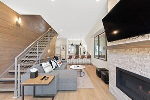Un salon moderne avec un canapé d’angle gris, une petite table basse, une cheminée et un coin cuisine à l’arrière-plan.