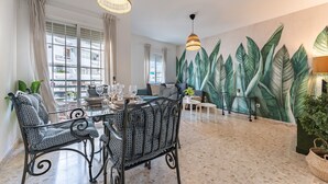 Un coin repas avec une table dressée pour quatre personnes, entouré de chaises à motifs, d’une grande fresque murale représentant des feuilles tropicales et offrant une vue sur la ville à travers de grandes fenêtres.