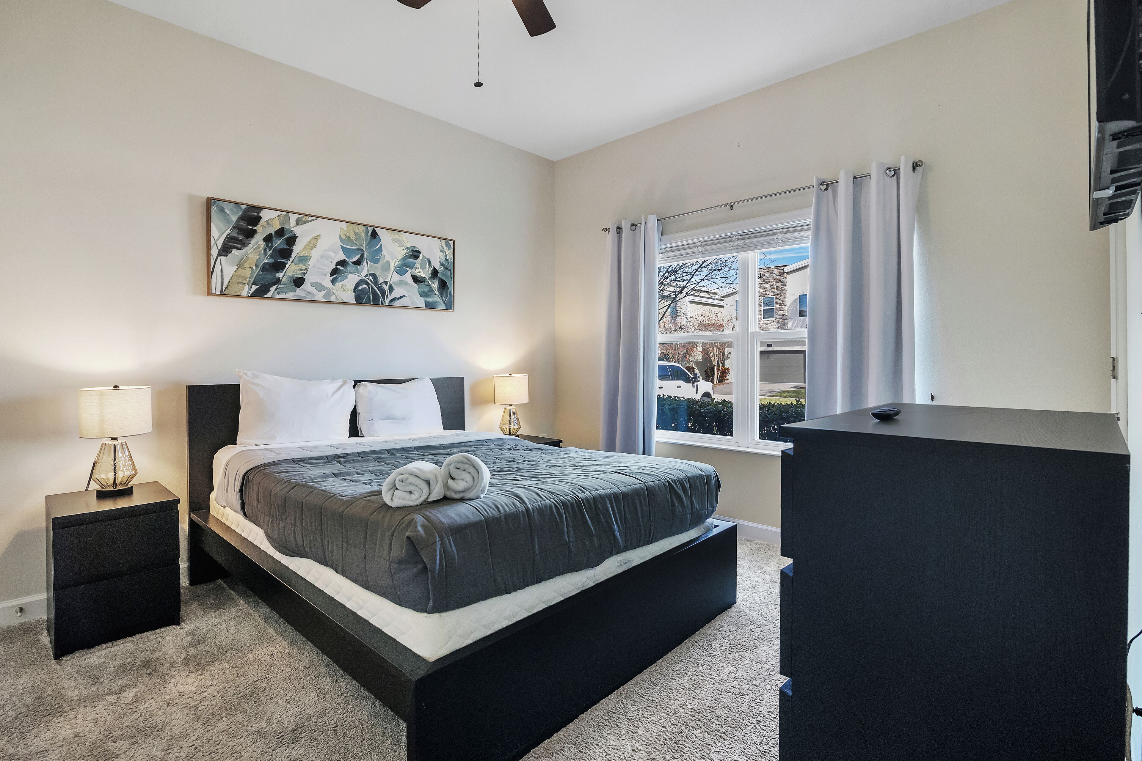A bedroom with a bed, nightstands, a desk, a window with curtains, and a painting on the wall.