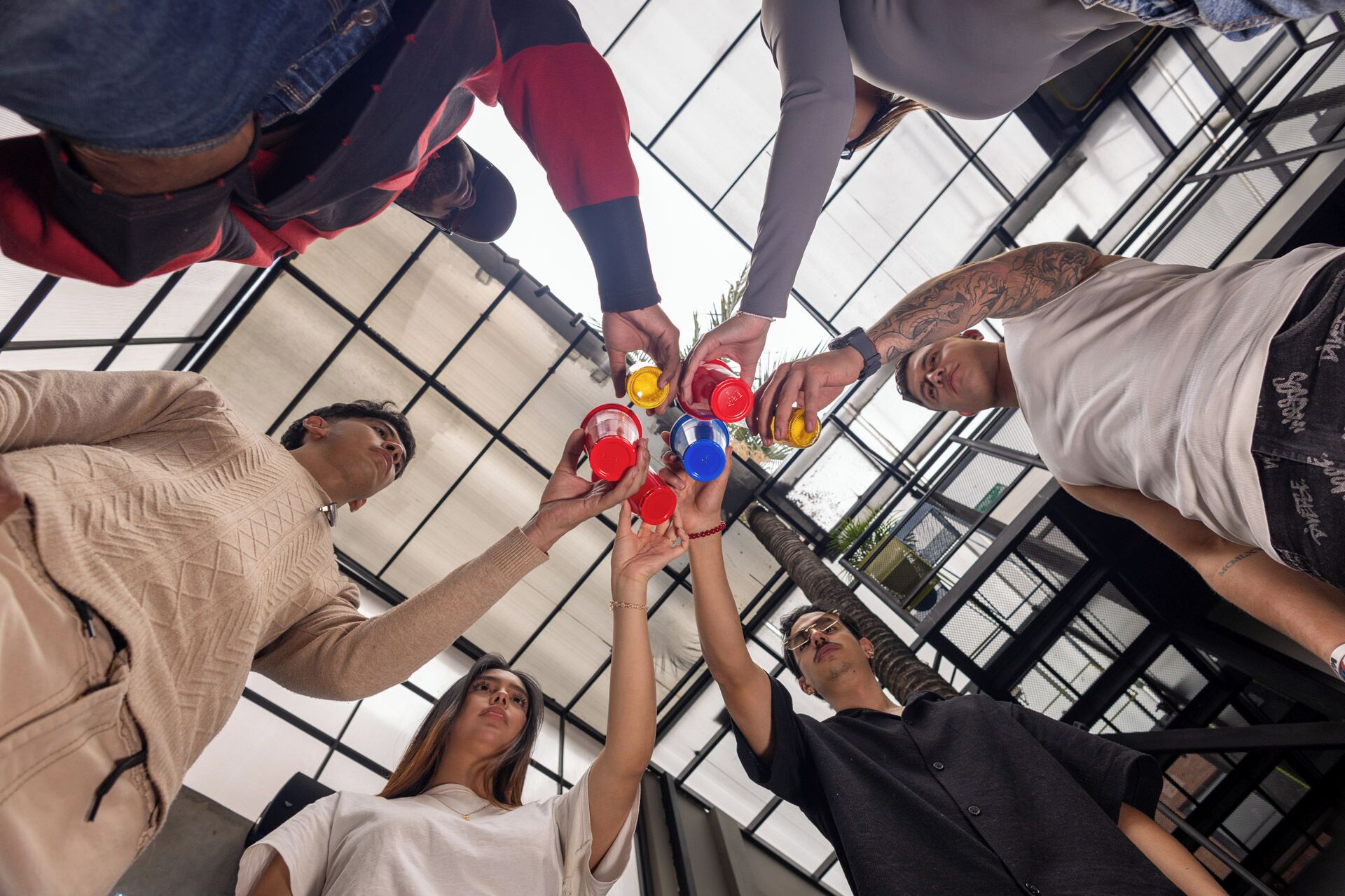 A group of people holding up colored cups in a high-ceilinged room with large windows.