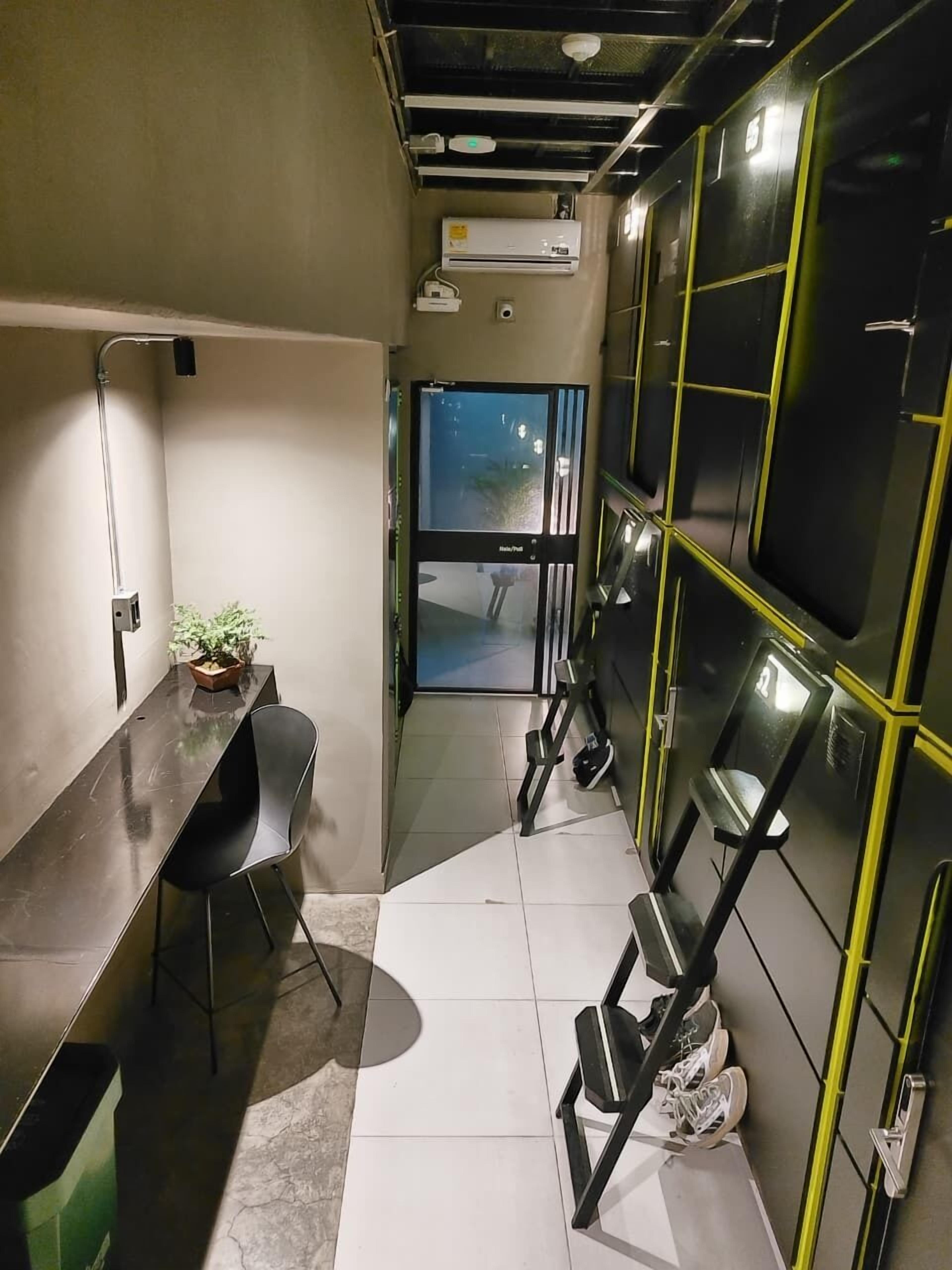 A narrow hallway with a desk, chairs, and a potted plant. There are black and yellow lockers along one wall and an air conditioning unit on the ceiling.