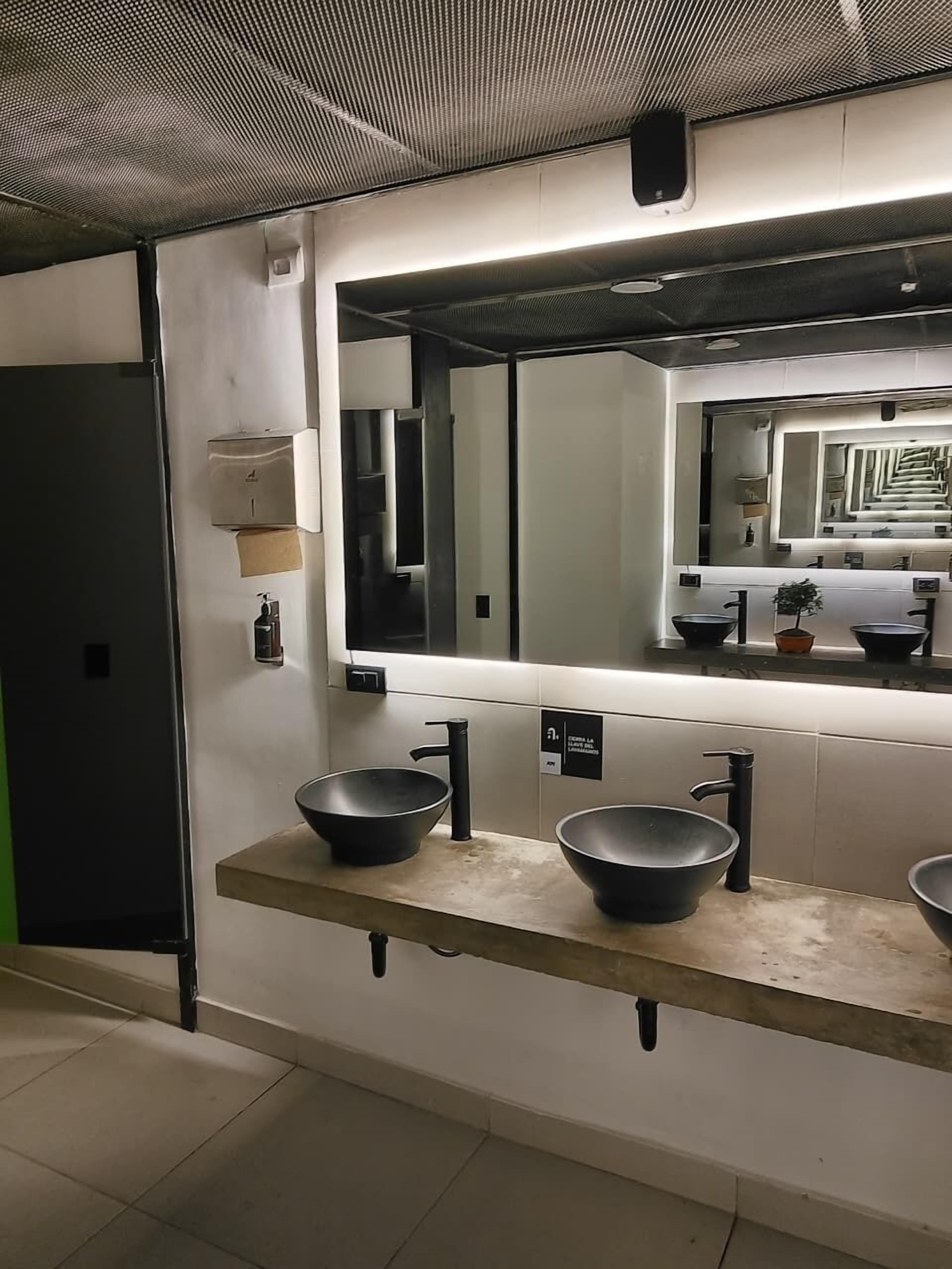 Un baño moderno con un espejo grande, dos lavamanos negros y un dispensador de toallas.