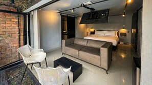 Une chambre d’hôtel moderne équipée d’un lit, d’un canapé et de fauteuils, d’une télévision et d’un balcon donnant sur un mur de briques.
