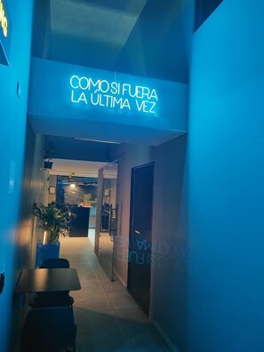 A hallway with a neon sign reading "COMOSI FUERA LA ULTIMA VEZ" and a reception desk in the background. - Hostal Joy Calle 85 (Bogotá)