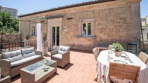 Un patio en la azotea con mobiliario de mimbre, una mesa de vidrio y una mesa de comedor con mantel floral.