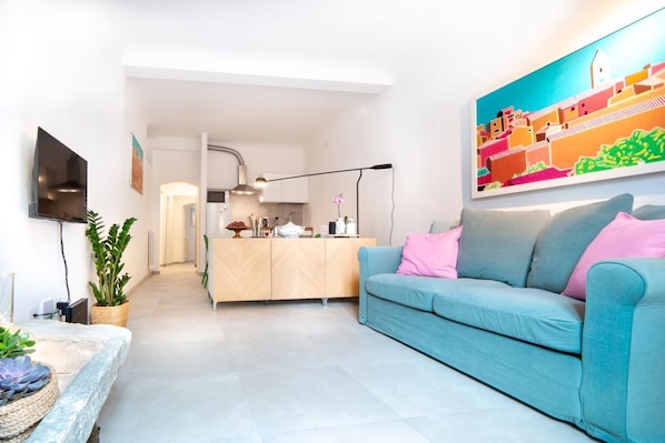 A modern living room with a sofa, a flat-screen TV, a kitchen in the background, and a colorful painting on the wall.