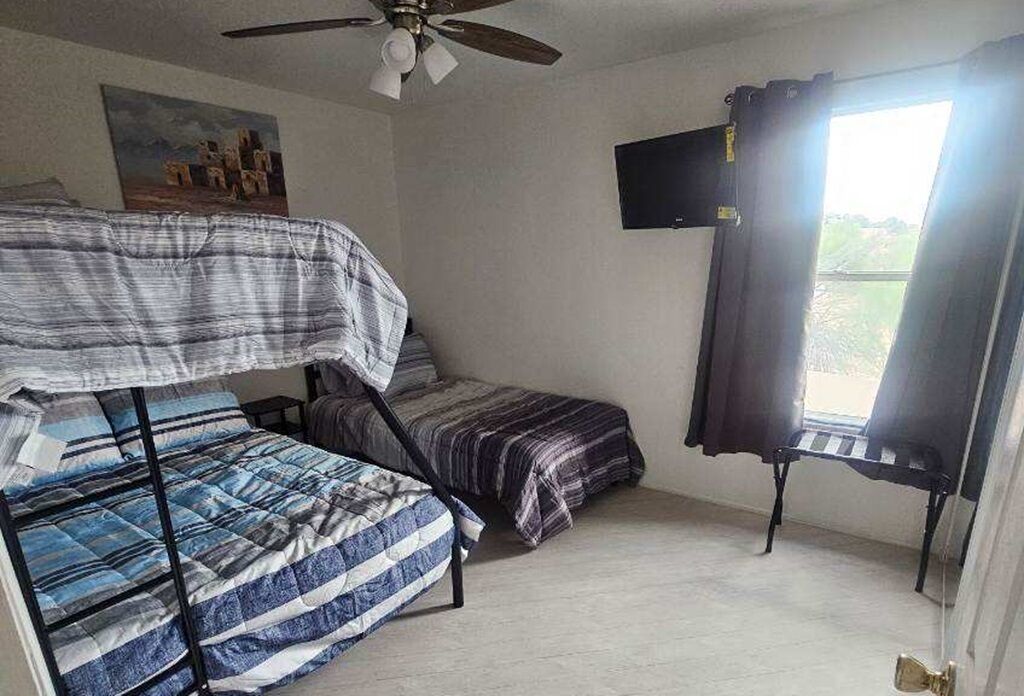 Quarto com beliche, uma cama de solteiro, ventilador de teto, televisão e janela com cortinas.
