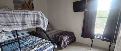 A room with a bunk bed, a single bed, a ceiling fan, a TV, and a window with curtains.
