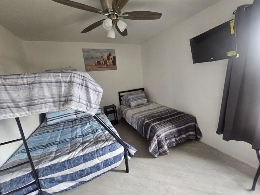 Een kamer in studentenresidentie met stapelbedden, een plafondventilator, een klein nachtkastje, een aan de muur bevestigde televisie en een schilderij.