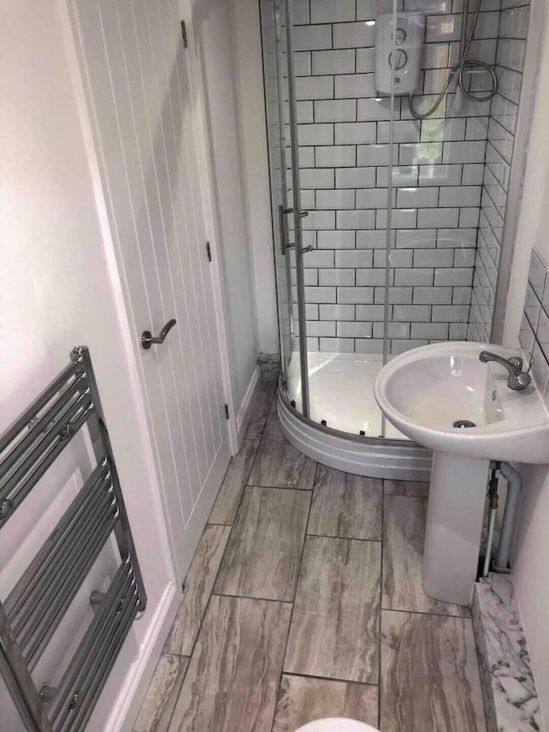 Un baño con cabina de ducha de vidrio, paredes con azulejos blancos, un lavamanos blanco y suelo de madera.