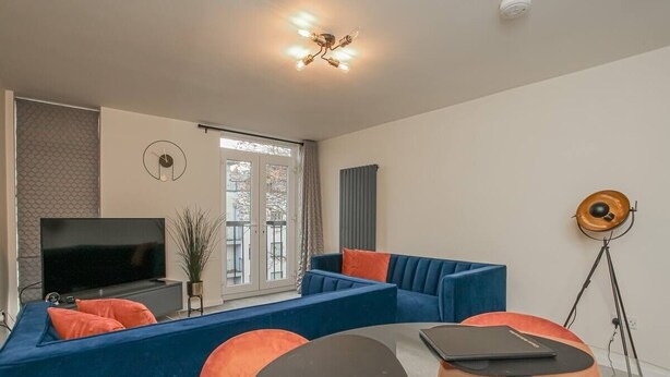 Een moderne woonkamer met een blauwe bank, oranje kussens, een ronde tafel met een laptop en een tv op een dressoir.