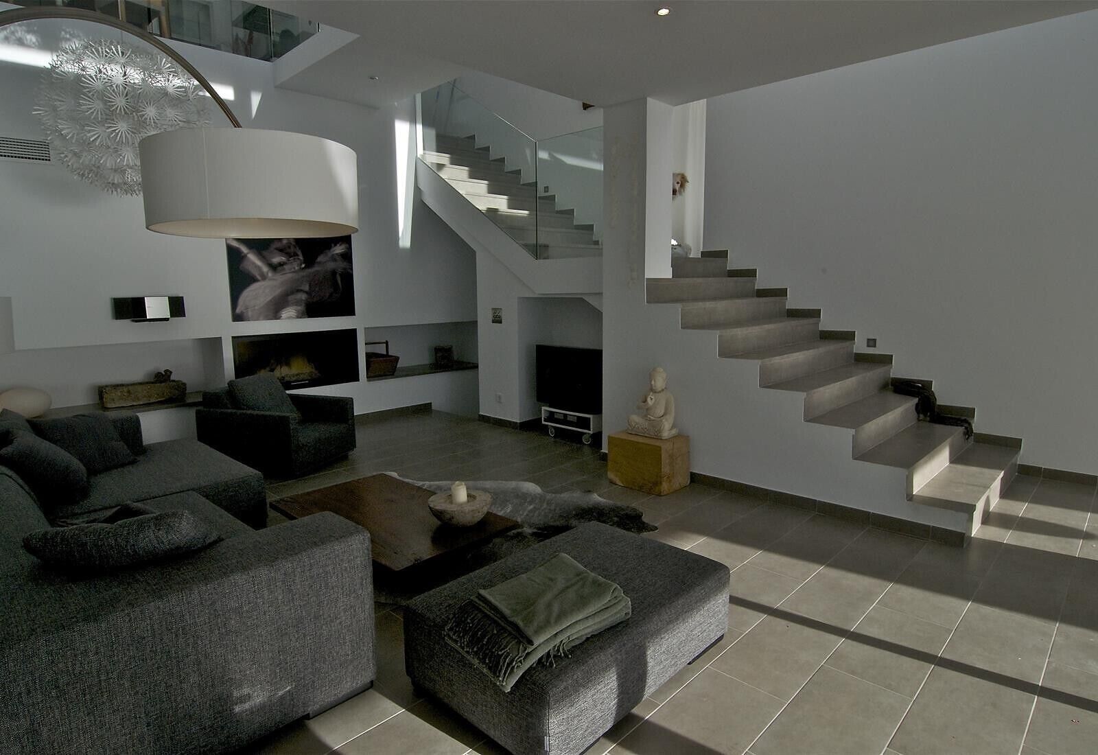A modern living room with a grey sofa set, a stone fireplace, and a staircase with glass railings.