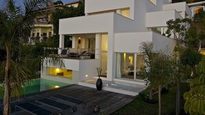 Una casa moderna de dos pisos con piscina, mobiliario exterior y abundante vegetación.