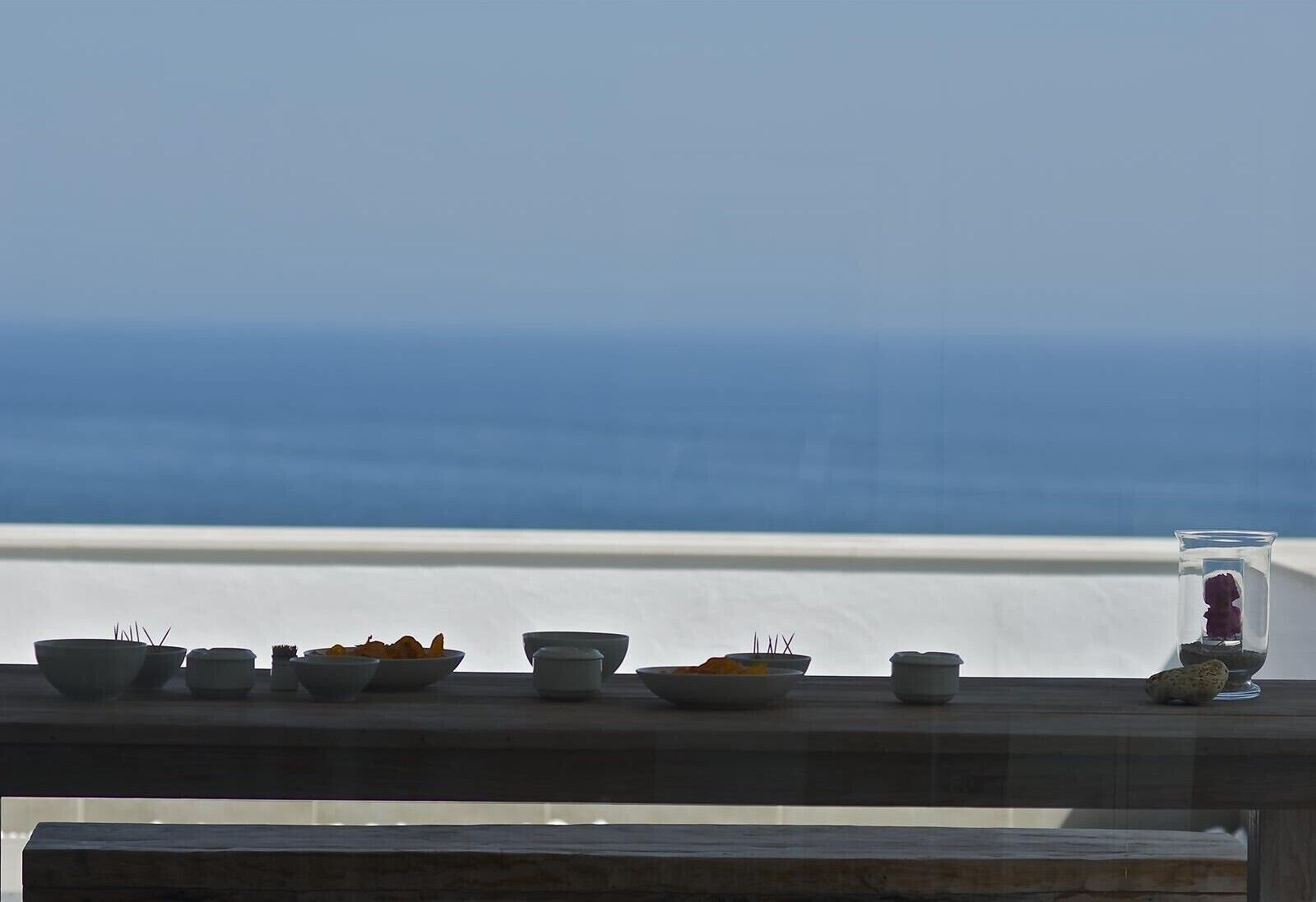 A balcony with a view of the sea, featuring a table with various dishes and a glass of water.