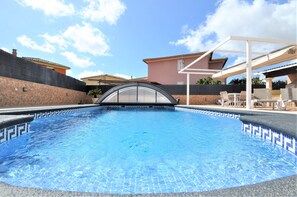 Una piscina con recinto in vetro, un patio coperto con sedie e un edificio sullo sfondo.
