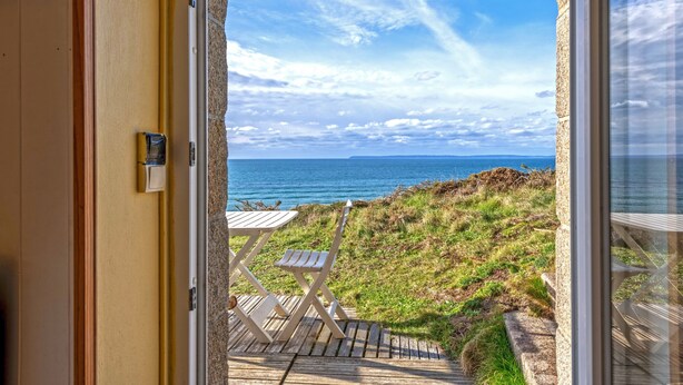Vue depuis une chambre dont la porte-fenêtre donne sur une terrasse avec une table et une chaise, d’où l’on aperçoit un espace verdoyant et la mer.