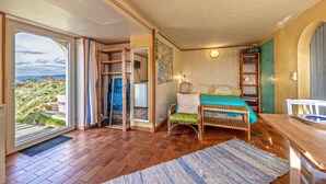 Une chambre avec une grande fenêtre, un plancher en bois, un lit, une chaise et une petite table.