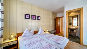 Hotellihuone, jossa on kaksi sänkyä, yöpöytä lampun kanssa, puinen penkki ja kylpyhuone, joka näkyy avoimesta ovesta.