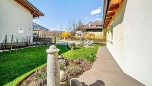 A residential courtyard with a stone pillar, a small garden, and a pathway leading to a house with a wooden roof. - Studio Apartment 'Apartment Typ C Haus Pöder' with Mountain View (Naturno)