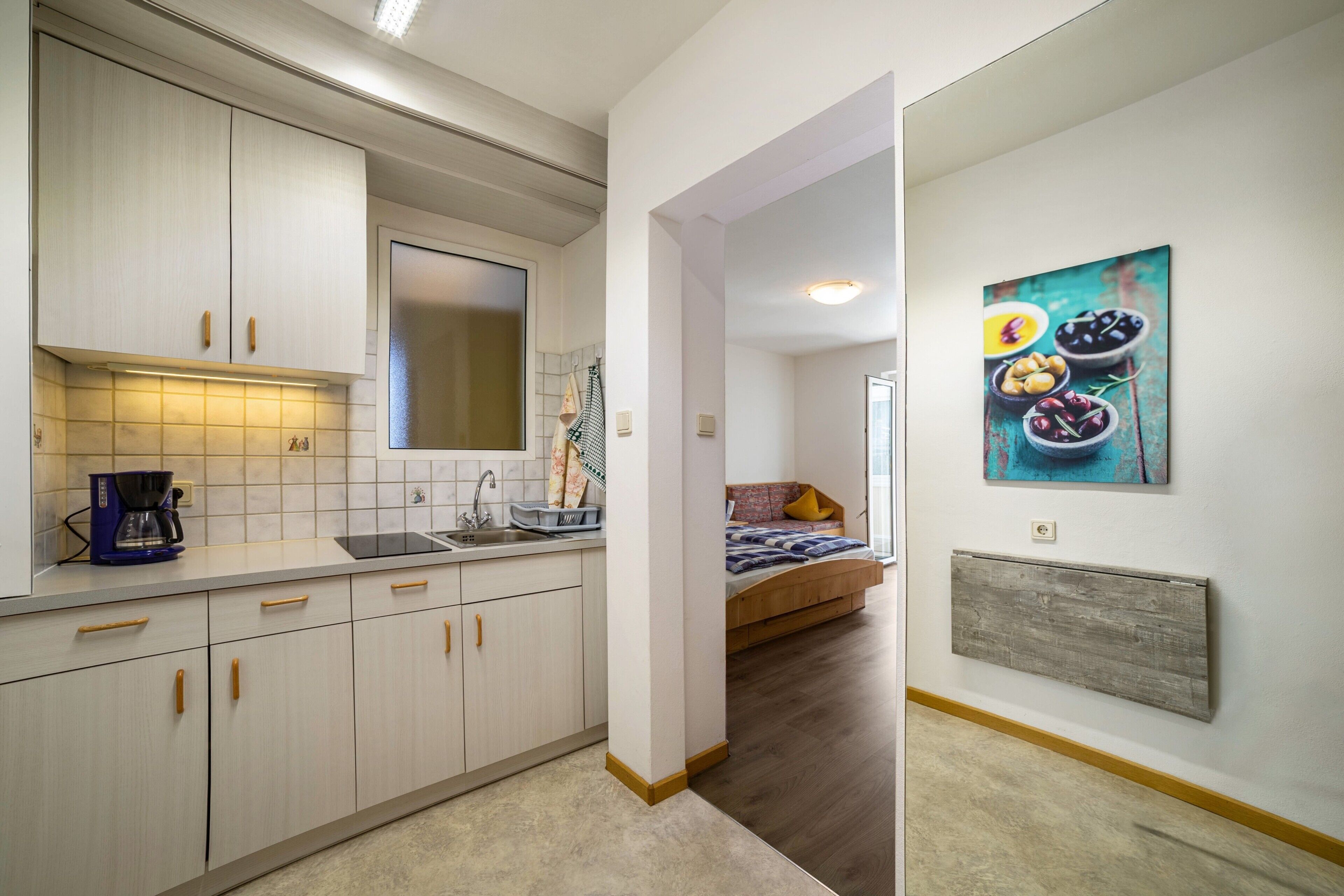 Eine moderne Küche mit weißen Schränken, einer Kaffeemaschine und einem Spülbecken. Im angrenzenden Zimmer befindet sich ein Bett mit einem blau-gelben Kissen.