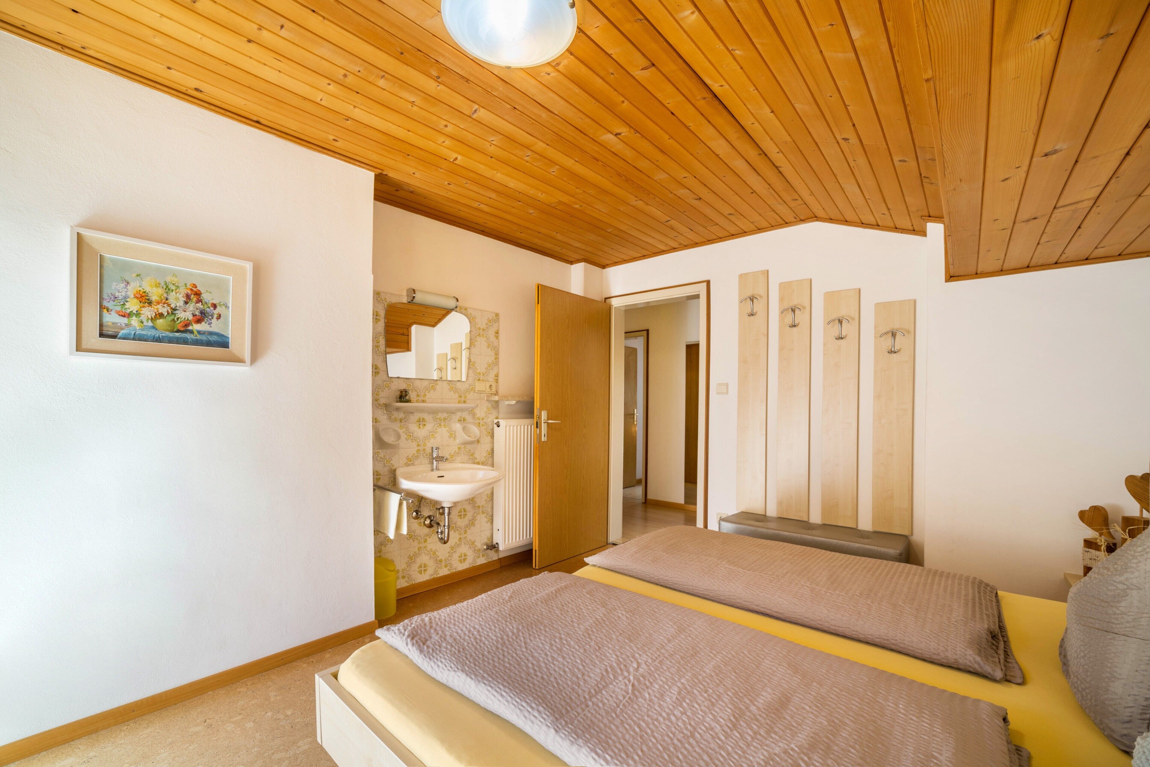 Un dormitorio con techo de madera, una cama, una mesita de noche, un espejo, un lavamanos y una puerta que da a otra habitación.