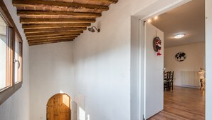 A hallway with a wooden door, a ceiling with exposed beams, a wall-mounted light fixture, and a view into a dining area with chairs and a table.