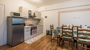A compact kitchen with a refrigerator, microwave, and stove, a dining area with wooden chairs and a table, and a small trash bin.