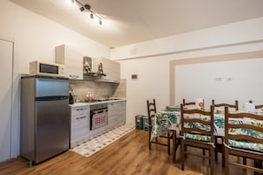 Une cuisine compacte équipée d’un réfrigérateur, d’un four à micro-ondes et d’une cuisinière, un coin repas avec des chaises et une table en bois, et une petite poubelle.