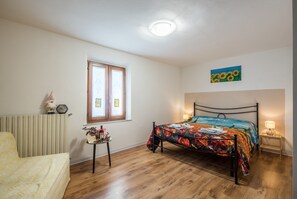 A bedroom with a bed, a window, a radiator, a small table with a plant, and a painting on the wall.