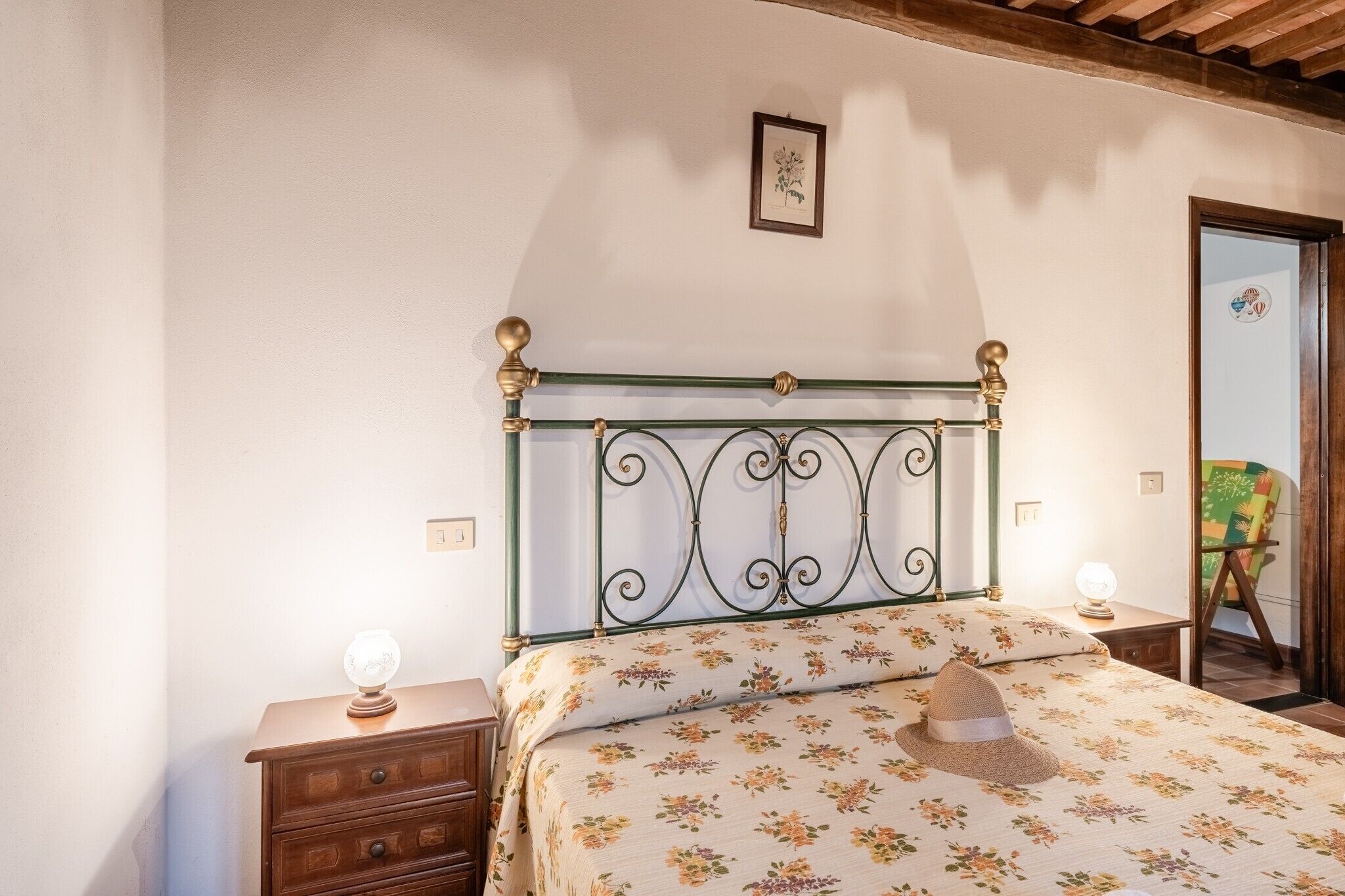 Une chambre à coucher avec une tête de lit en métal, une literie à motifs floraux, une table de chevet en bois avec une lampe, et une porte donnant sur une autre pièce.