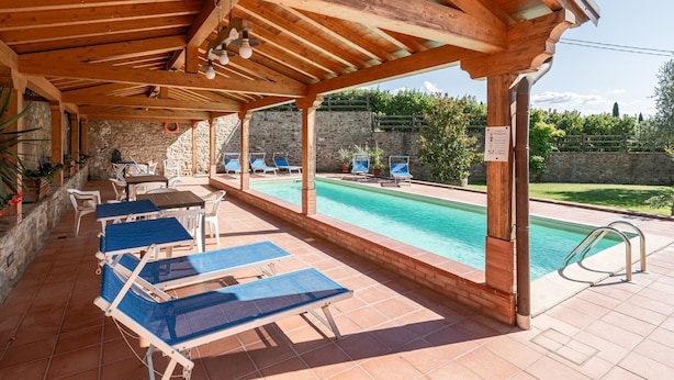 Zona de piscina al aire libre con pérgolas de madera, sillas de descanso y un muro de piedra de fondo.