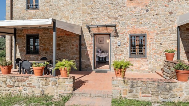 Un edificio de piedra con un espacio de estar techado al aire libre, una puerta que da a un interior y plantas en macetas.