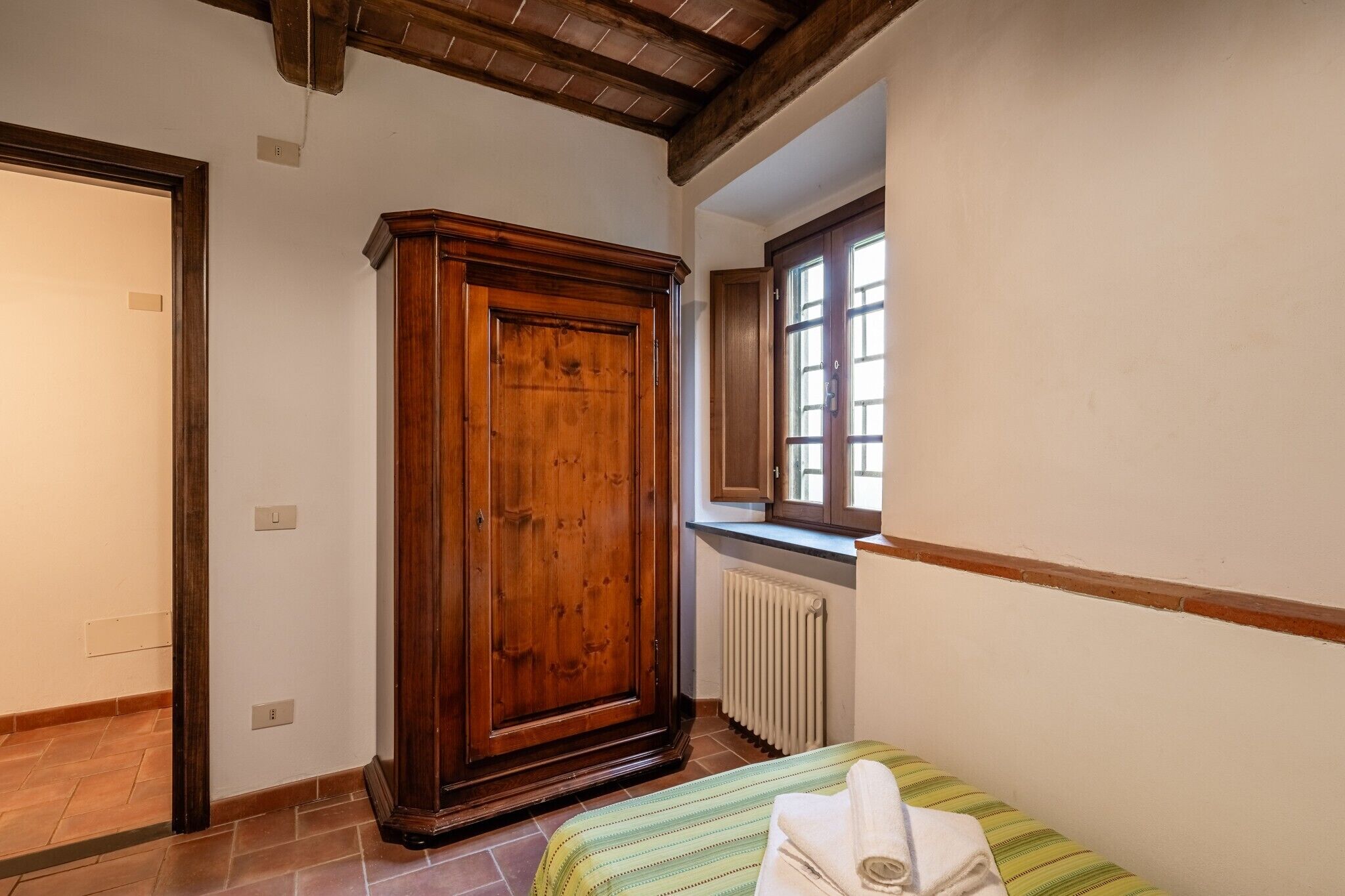 Ein Schlafzimmer mit Holzschrank, Heizkörper, einem Fenster mit Jalousien und einem Bett mit gestreifter Bettdecke.