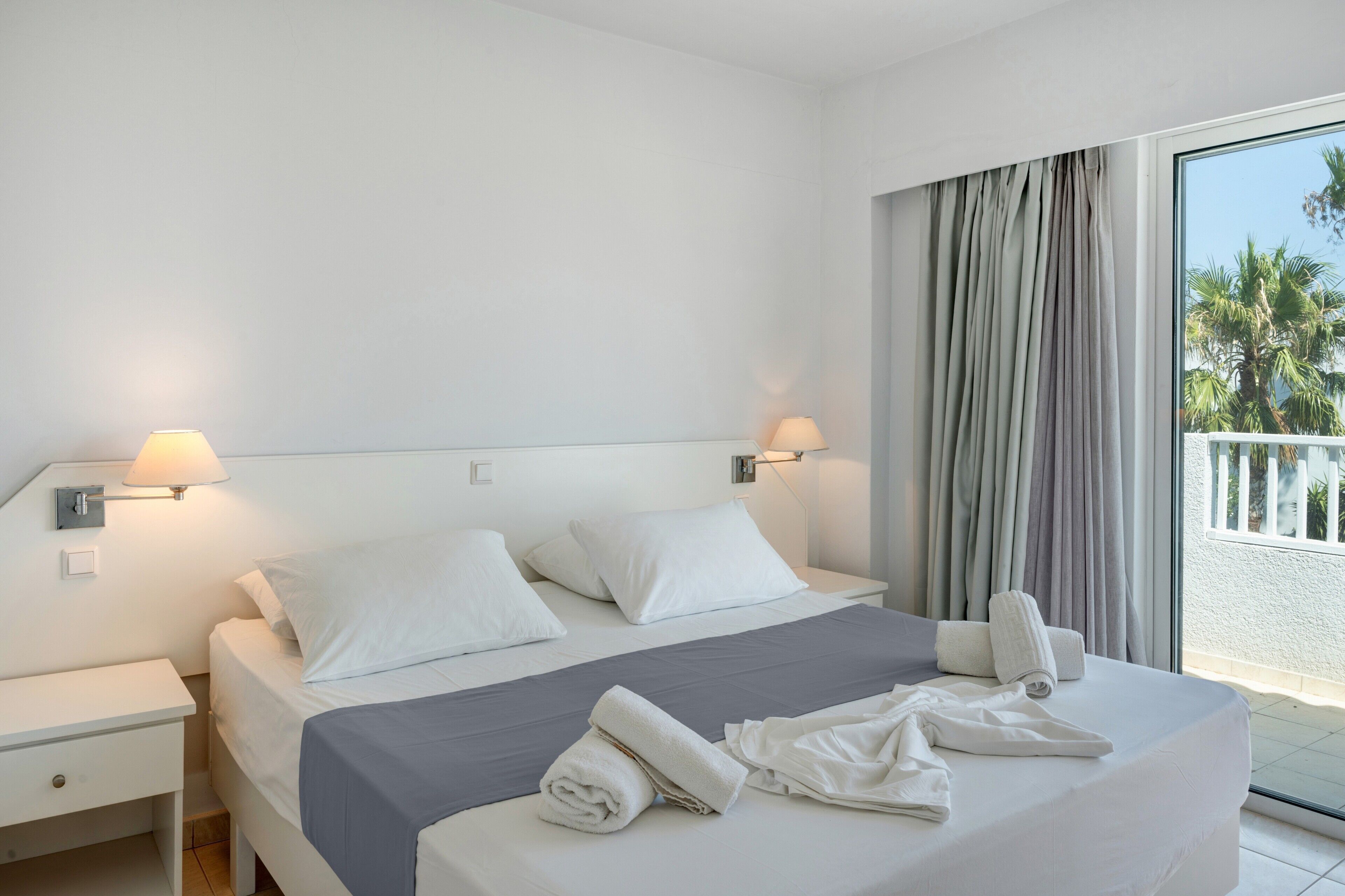 Habitación de hotel con cama, dos mesitas de noche, ventana con cortinas y vistas a palmeras.