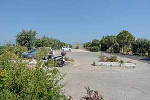 A paved driveway leading to a building with a stone facade, surrounded by greenery and parked vehicles.