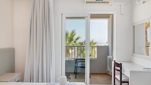A hotel room with a bed, a desk, a chair, a balcony with a view of palm trees and the sea, and an air conditioning unit. - Studio Apartment 'Evripides Triple Room 2' with Shared Pool, Shared Garden and Air Conditioning (Kardamaina)